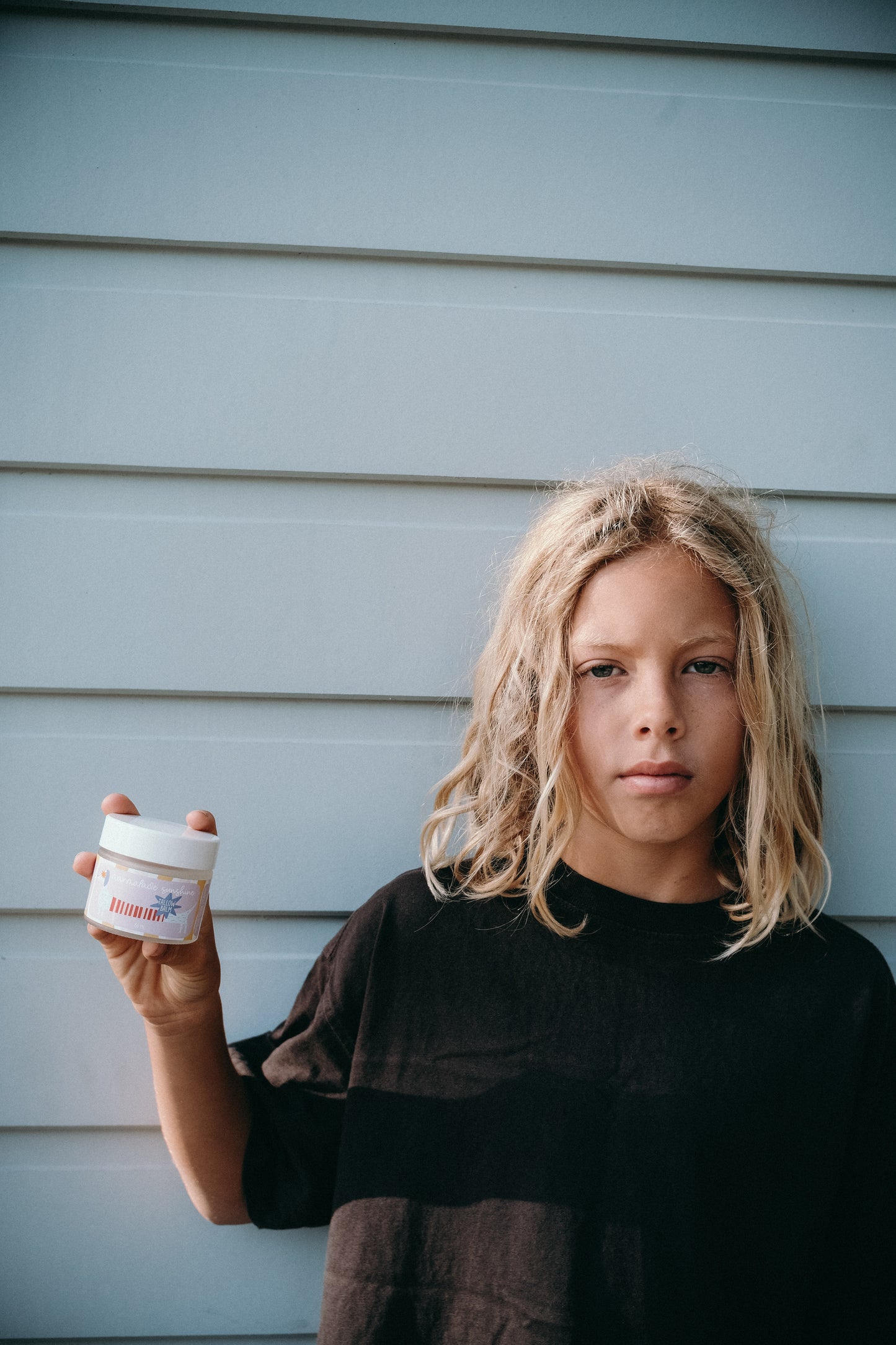 Child holding Glass Wholefoods Tallow Balm jar in front of a light blue weatherboard wall