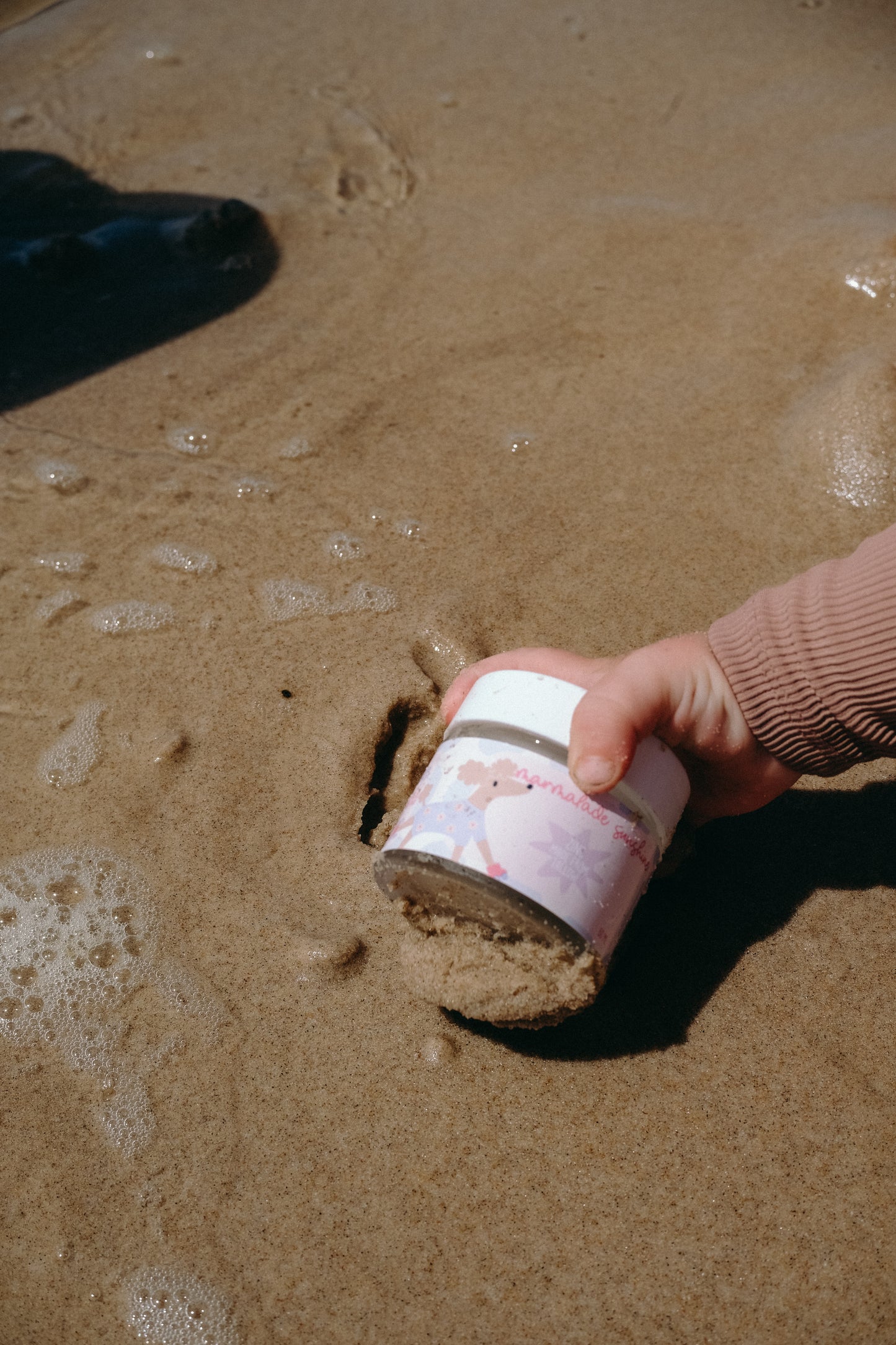 Hand reaching for Glass Wholefoods Tallow Zinc Sun Balm jar on wet sandy beach