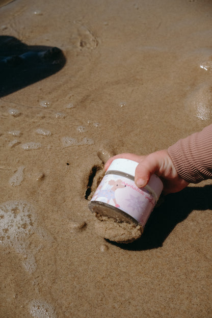 Hand reaching for Glass Wholefoods Tallow Zinc Sun Balm jar on wet sandy beach