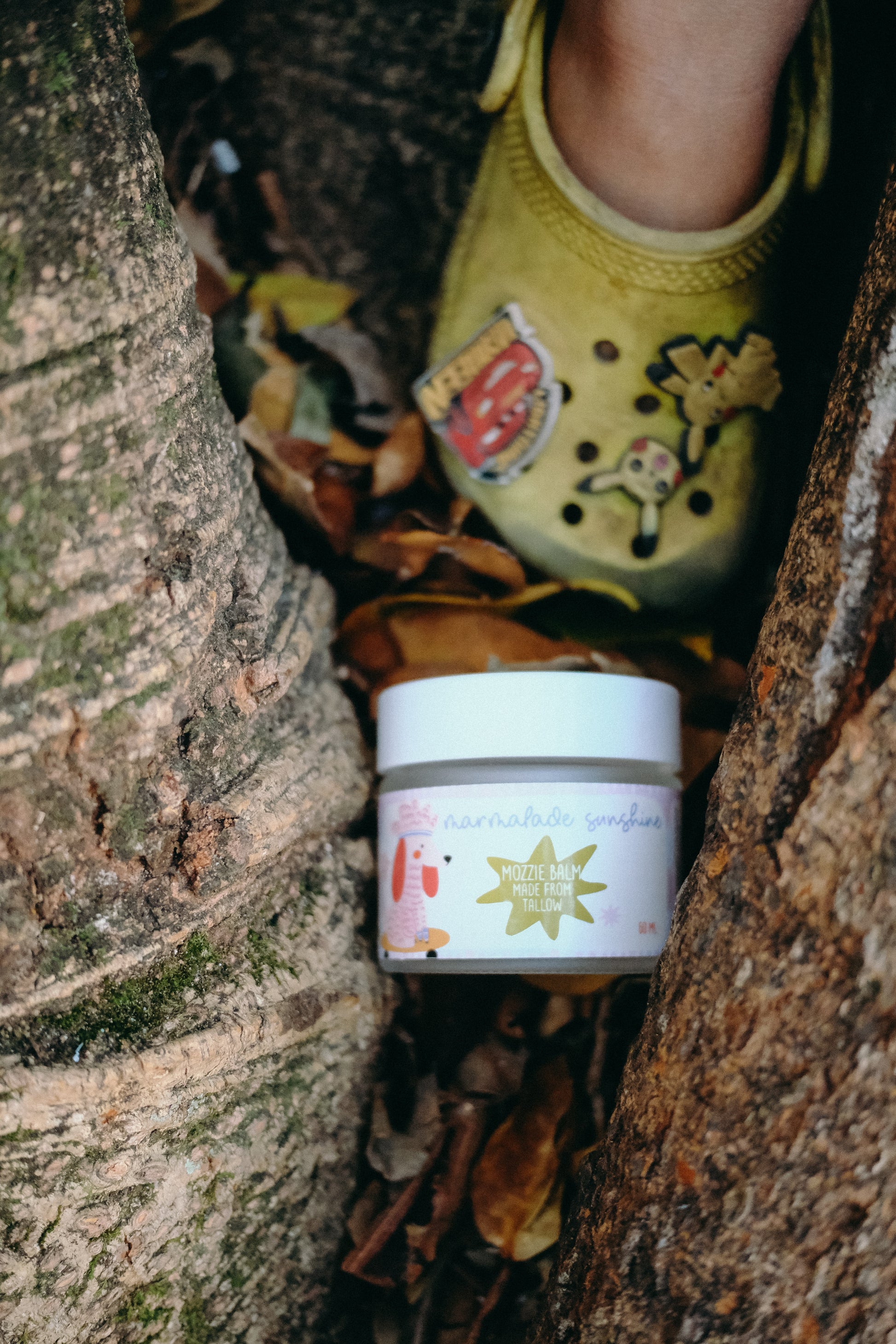Glass Wholefoods Tallow Mosquito Balm jar nestled in tree bark outdoors with child's feet above